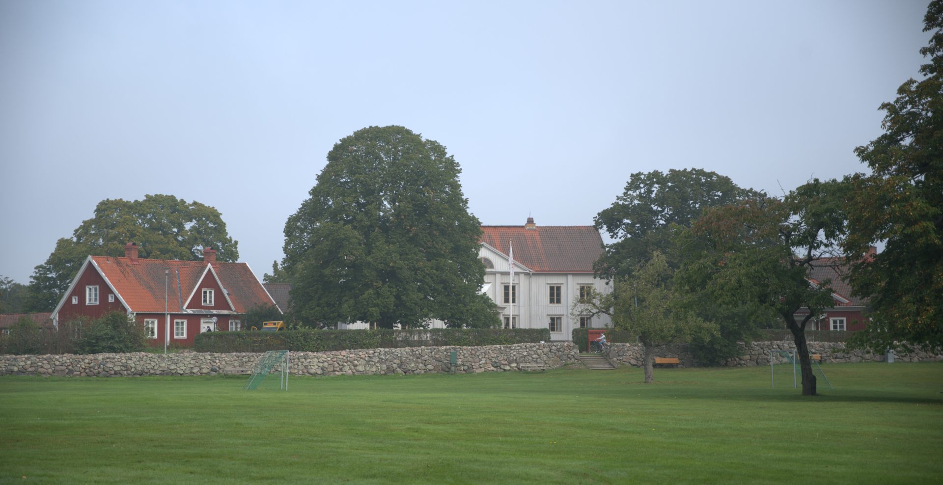 Skälby Kungsgård med stora träkronor och en stenmur i förgrunden. Ett vitt stort trähus i mitten. Till vänster är ett rött, lite mindre trähus. Den västra flygeln. 