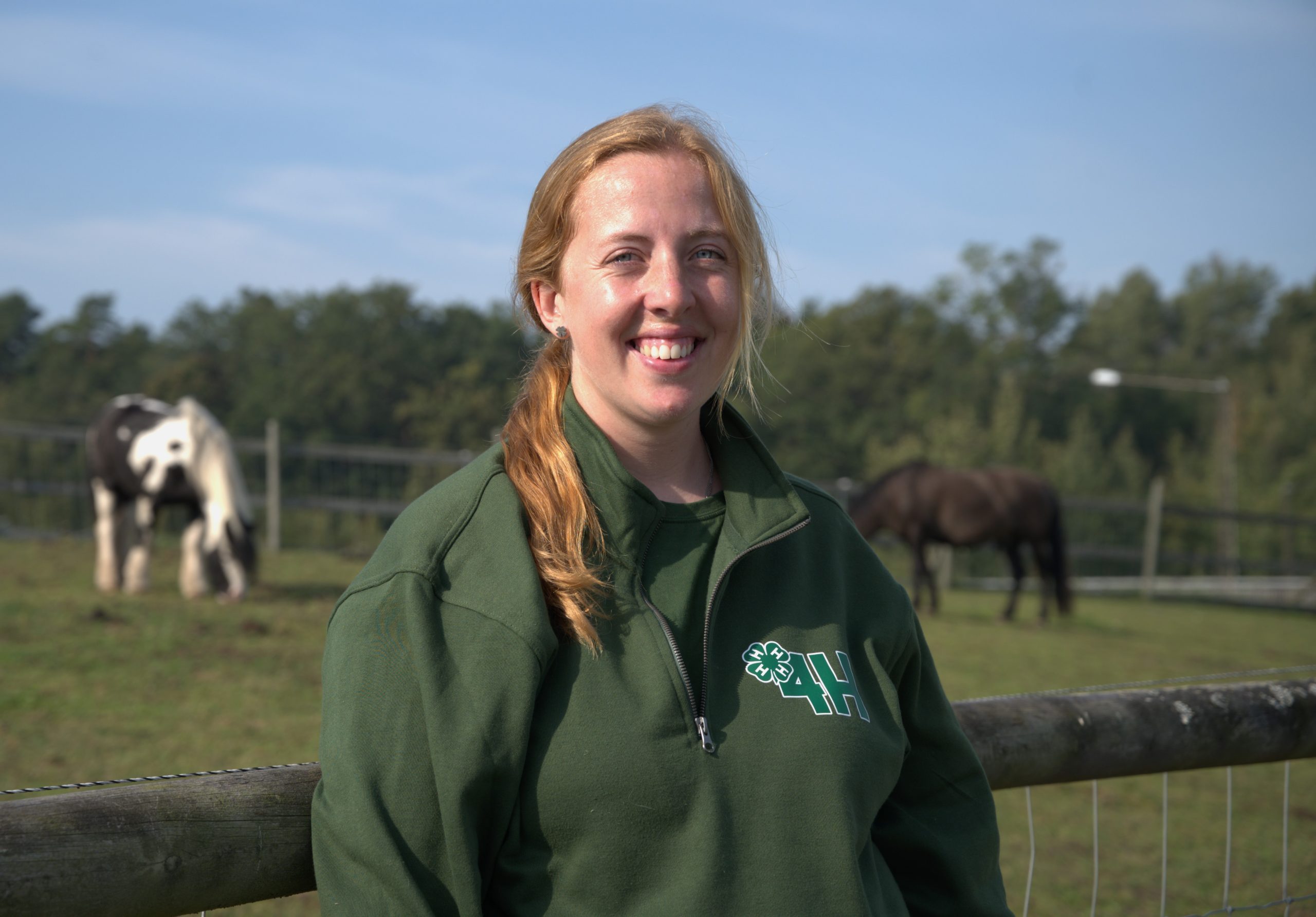 Isabelle Hall Wilkens, 32, framför en hästhage. Bakom sig till vänster i bild är en brunvit häst. Till höger i bild står en gråsvart häst. 