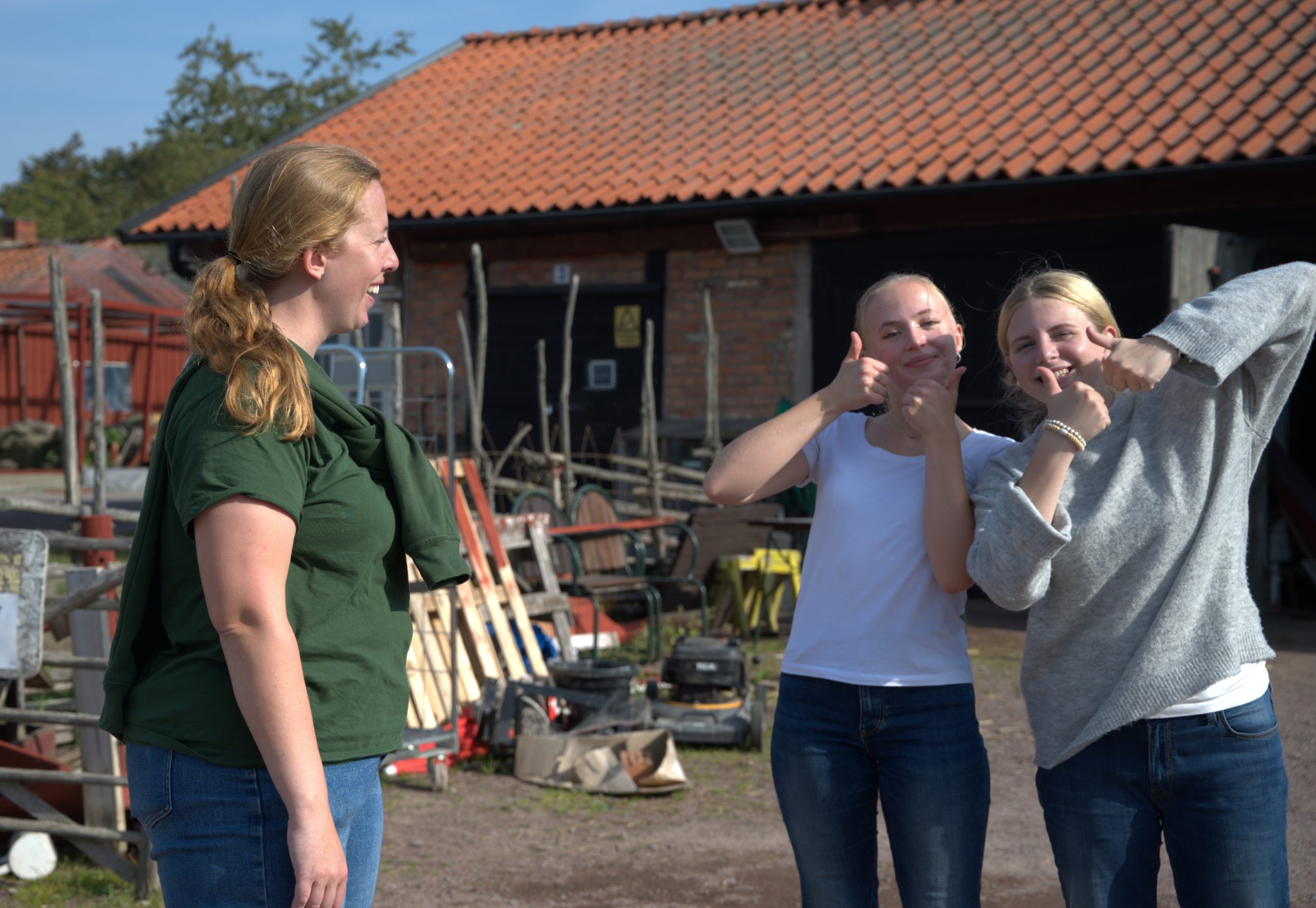 Isabelle står och pratar med två tjejer som är aktiva på gården. De tittar in i kameran och ler och håller upp sina tummar. Isabelle tittar på och ler.