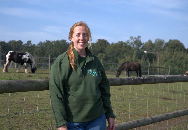 Porträttbild av Isabelle Hall Wilkens framför en hästhage. Till vänster i bild är en vit och svartfläckig häst och till höger står en gråsvart.