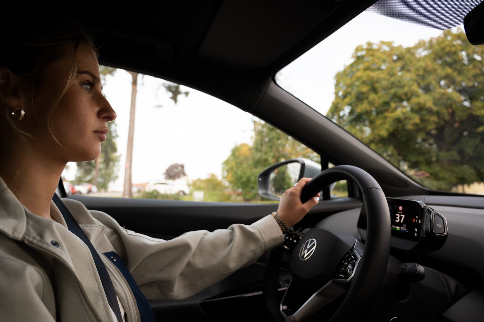 Andrea sitter I förarsätet i bilen och håller ena handen på ratten. Det är träd i bakgrunden. Andrea kollar rakt fram och kör bilen.