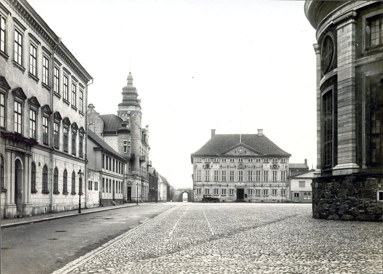 Fotografi på ett torg med en väg och byggnader i bakgrunden.