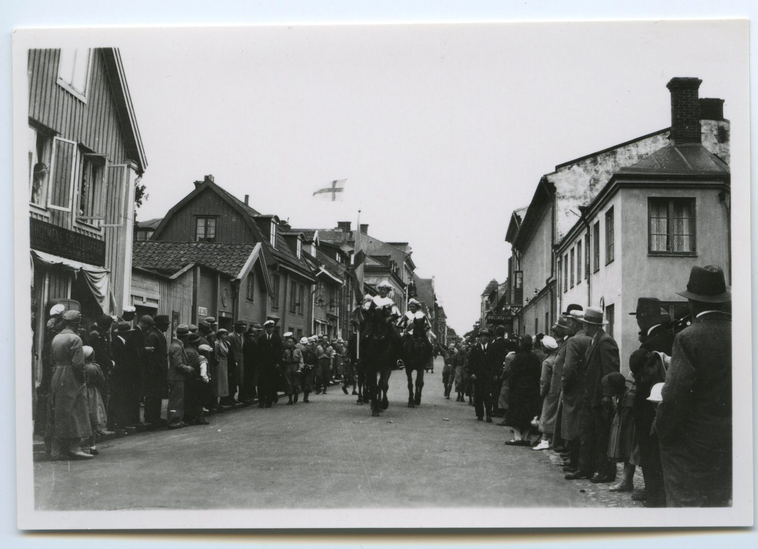 En bild på en gata, hästar går i mitten och folk står och tittar på vid sidorna.