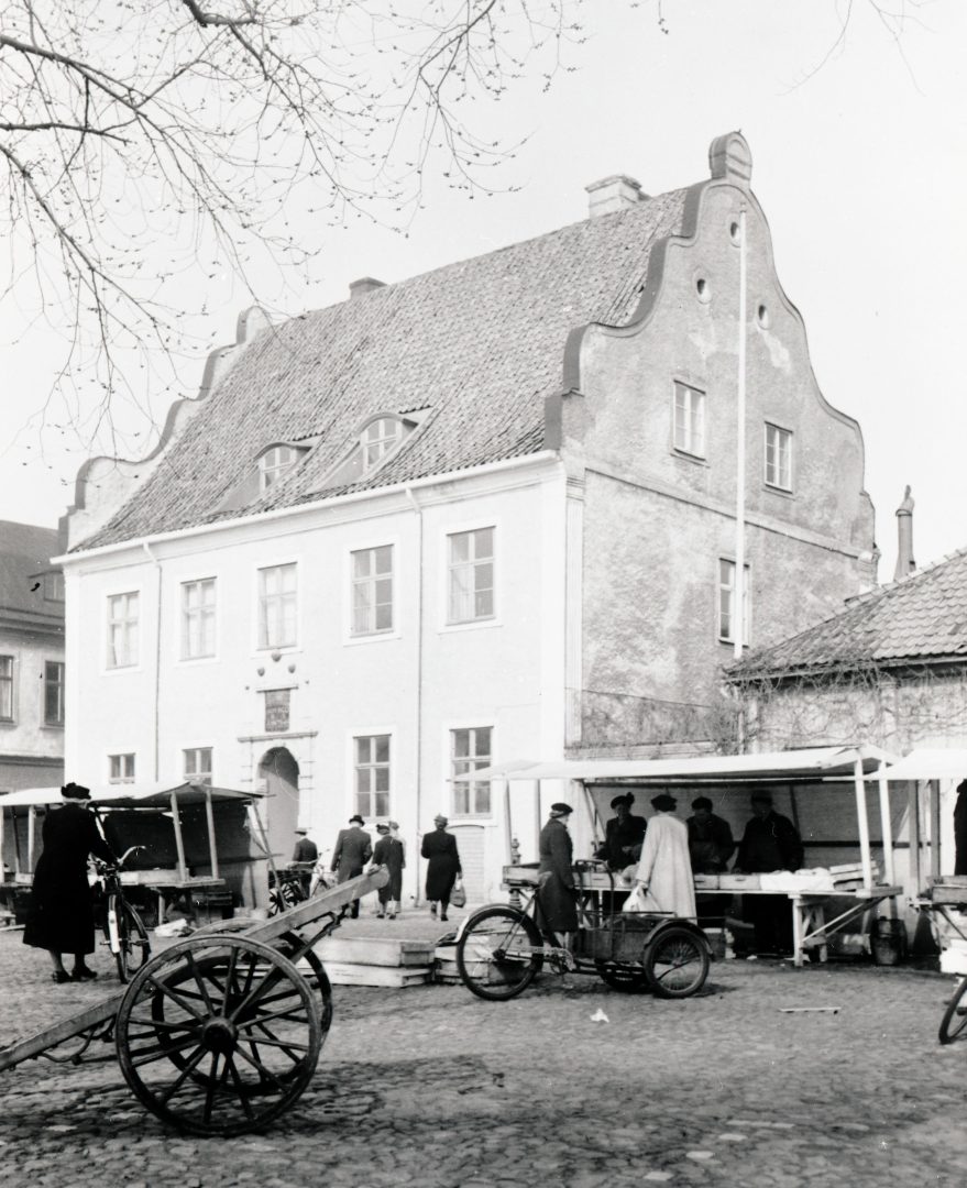 Ett svart-vitt foto på ett hus och ett torg. Människor handlar från en marknad.