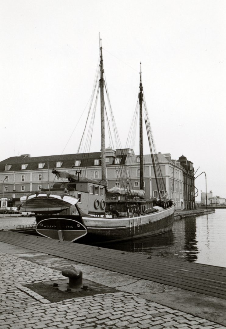 Fotografi på en segelbåt som lagt till i en av Kalmars hamnar, bakom ser man ett hus.