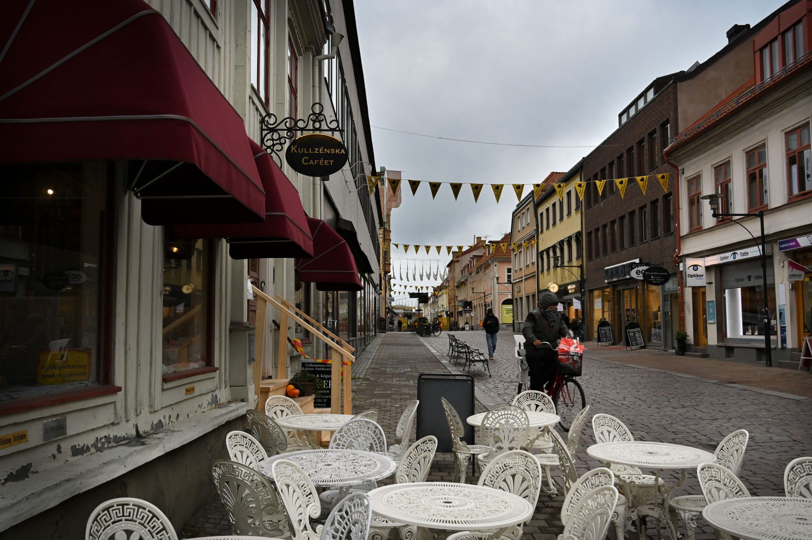 En bild på en gågata med en uteservering till vänster i bilden. En dam kommer cyklandes, och en man går längre bak.