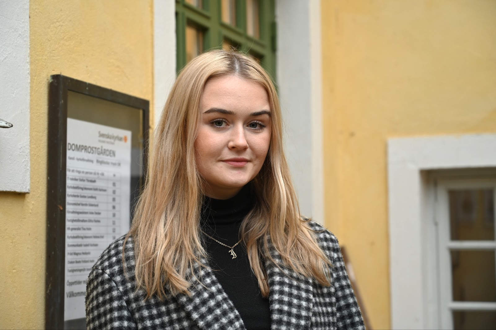 Isabell Karlsson är kommunikatör för Svenska kyrkan i Kalmar. Hon står framför en gul husfasad och en grön dörr. Isabell har blont hår, en svart tröja och en rutig jacka på sig. 