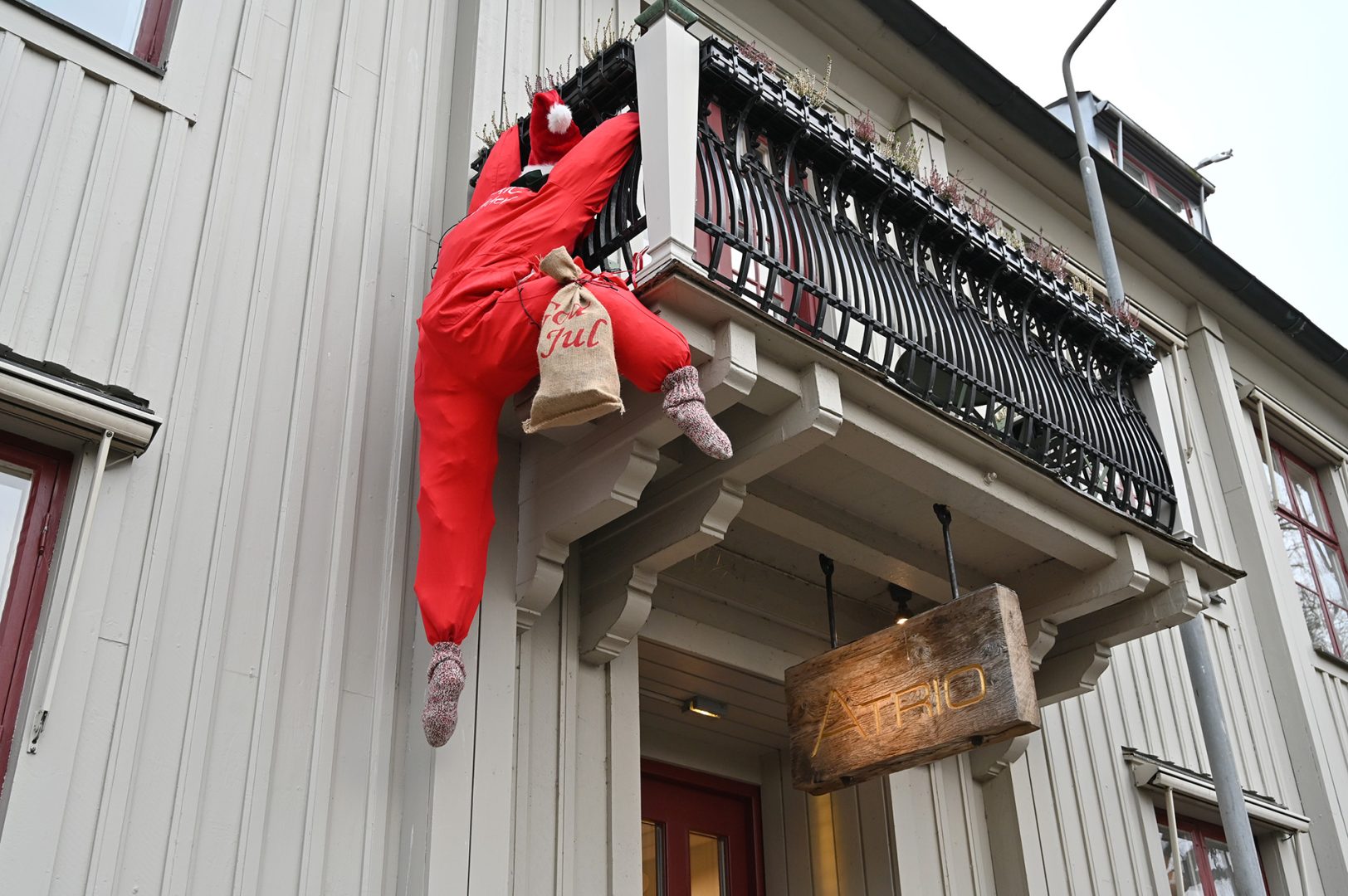 En låtsastomte som håller i sig i ett balkongräcke. En juldekoration där det ska se ut som att tomten håller fast sig i balkongräcket. Tomten har på sig en röd tomtedräkt.