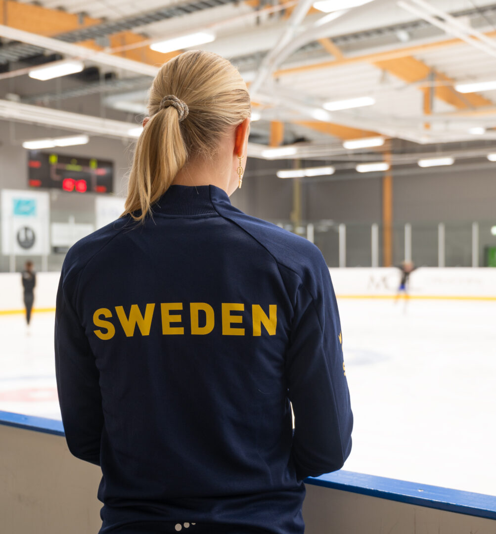 Alice kollar ut över isen där ett par barn åker runt. På hennes blå träningströja står det Sweden i gult på ryggen. 