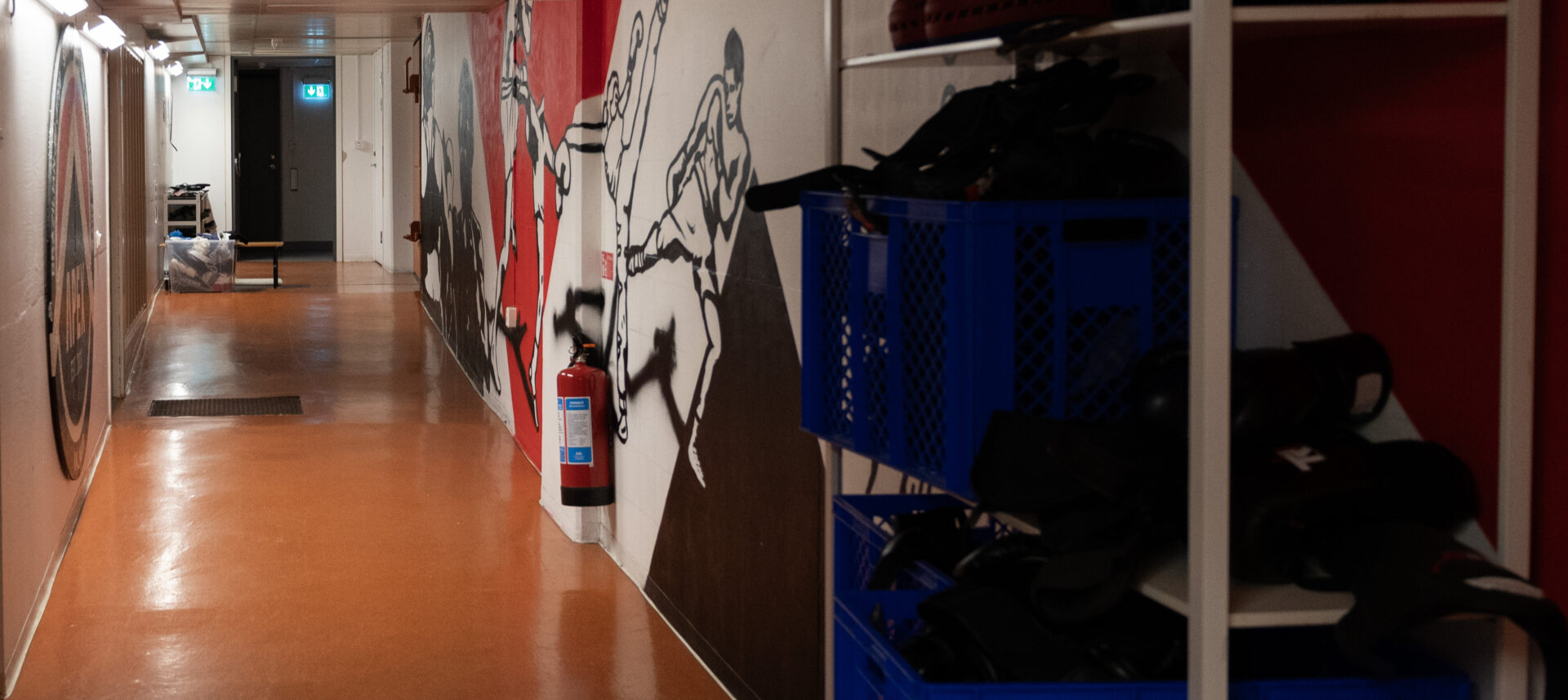 Korridor påväg in i klubben. Golvet är oranget och väggen är täck med ett konstverk av folk som utför olikakamproter. I höger kant av bilden står det en hylla med mitsar och handskar som används under thaiboxnings träningen