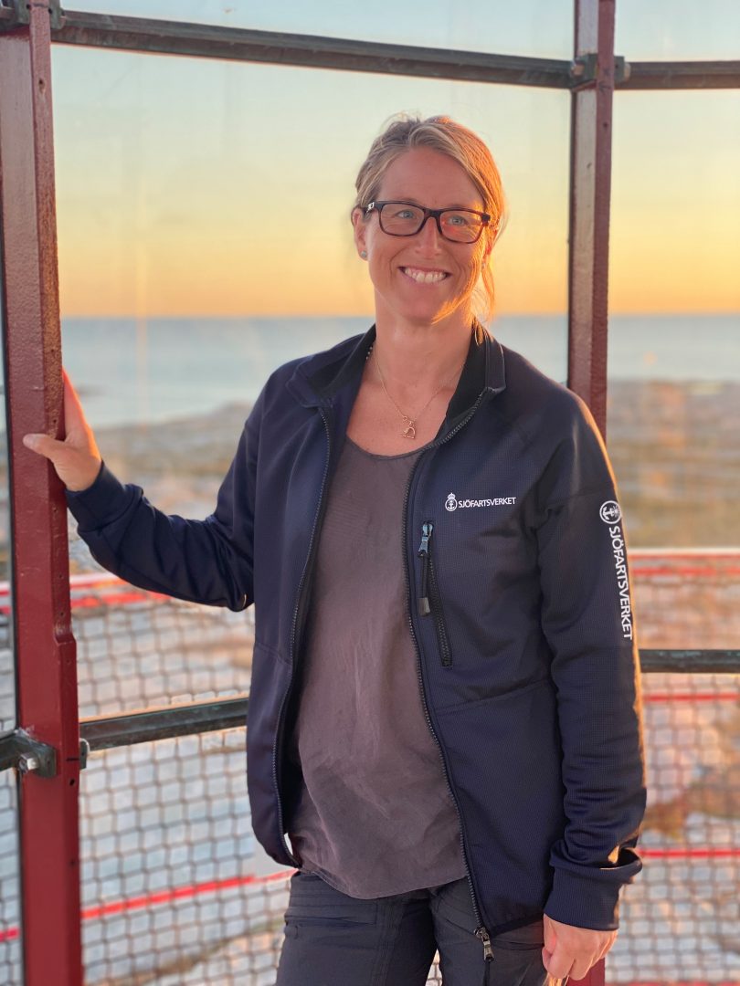 En kvinna står i ett fyrtorn vid solnedgången. Hon ler mot kameran och har glasögon samt en mörk jacka med Sjöfartsverkets logotyp på bröstet och ärmen. I bakgrunden syns havet och himlen som färgats orange av kvällssolen.