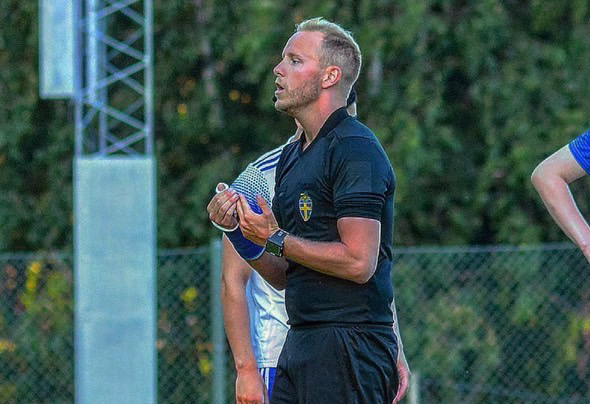 En fotbollsdomare står utomhus på en plan med gräs och träd i bakgrunden. Han bär en svart domartröja och håller en liten anteckningsbok eller kortblock i händerna. Hans blick är riktad åt höger, som om han följer spelet eller talar till någon utanför bild. Bakom honom syns spelare i blått och vitt.