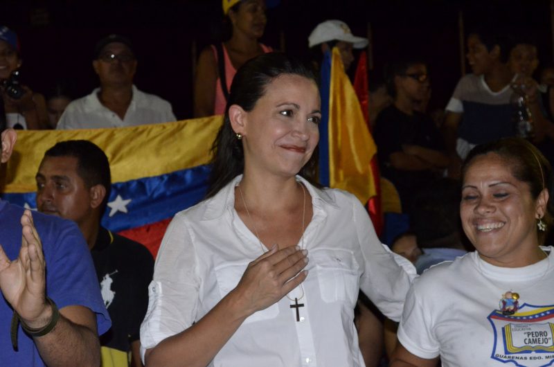 María Corina Machado framför Venezuelas flagga. Hon håller handen mot bröstet. Runt halsen har hon ett halsband med ett kors.
