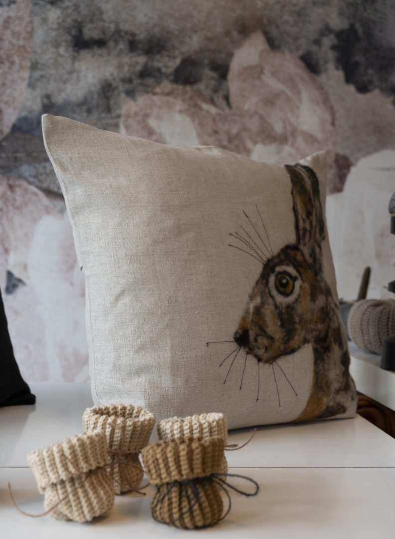 På ett bord syns en beige kudde med en hare på, framför den står fyra små stickade strumpor.