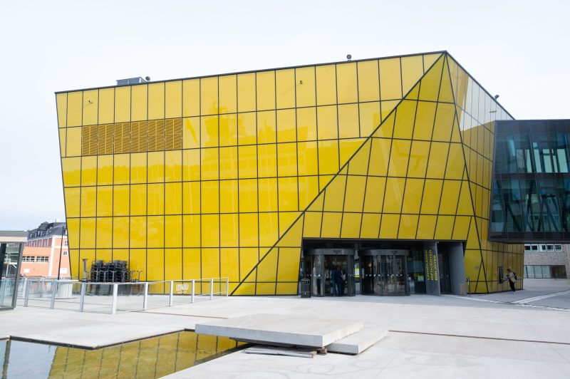 En modern byggnad med fasad av gula glasrutor och svarta linjer som bildar geometriska mönster. Entrén har glasdörrar, och till höger finns en del av byggnaden med blåtonade glasrutor. Framför byggnaden syns en betongyta och en liten vattenspegel.