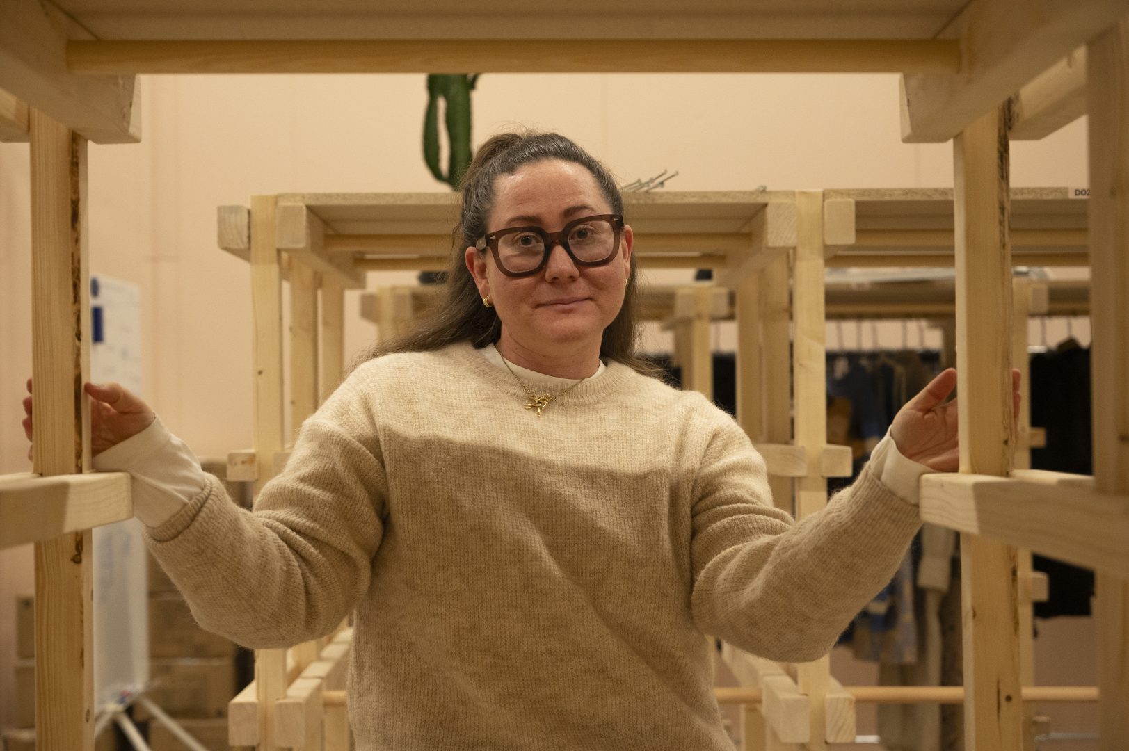 En 30-årig kvinna med mörkt hår och stora glasögon står i en trähyllla. Hon bär en beige stickad tröja. Hon har ett neutralt ansiktsuttryck. I bakgrunden ser man fler trähyllor där det hänger kläder.