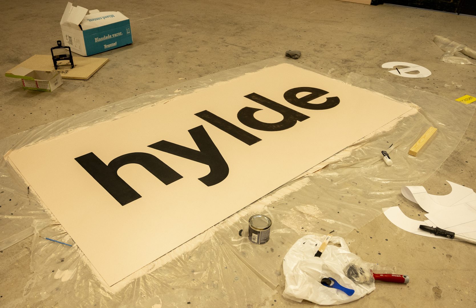 En ljusrosa skylt ligger på ett genomskinlig plastskynke. På skylten står det: hylde. Omkring skylten ligger det penslar och burkar med färg.