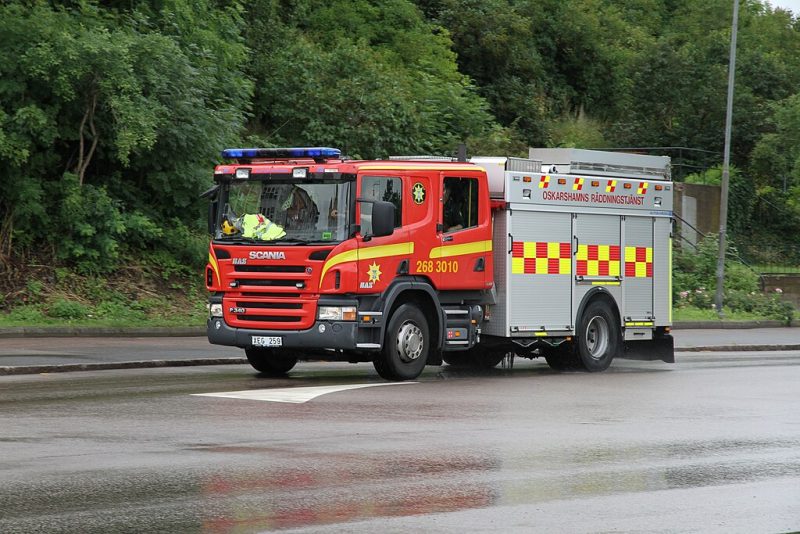 En bild på en rödgul brandbil som kör på en blöt väg. I bakgrunden sysn gröna träd.