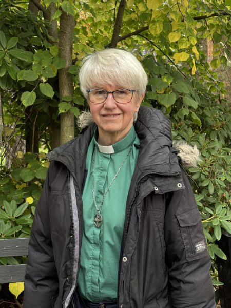 En äldre person med kort, grått hår och glasögon står utomhus framför grönskande buskar och träd. Personen bär en svart jacka med pälsfodrad huva och en grön prästskjorta med vit krage, samt ett halsband med ett kors.