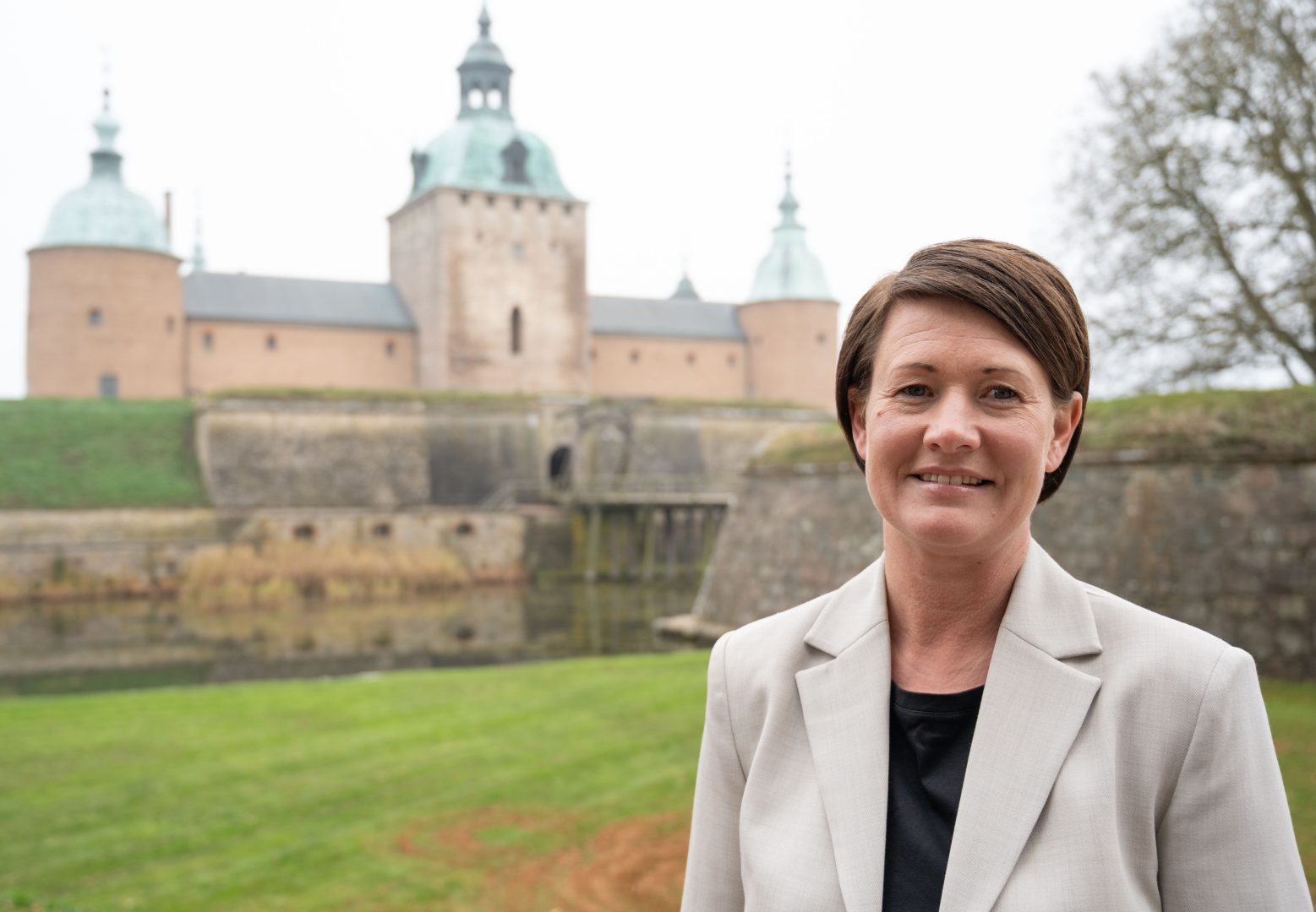 En kvinna med kort brunt hår, klädd i en ljusbeige kavaj och svart topp, står leende utomhus framför en historisk borg med torn och gröna kopparkupoler. Gräsmatta och träd syns i bakgrunden under en molnig himmel.