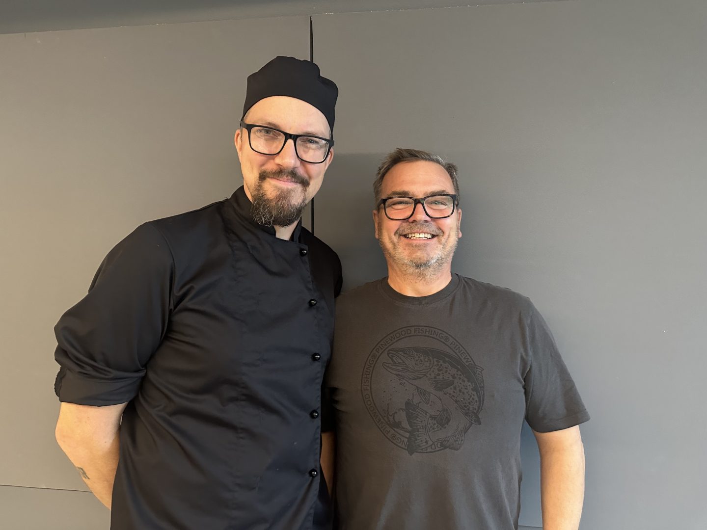 Två män står bredvid varandra och ler. De står mot en grå vägg. Den ena mannen har på sig kock kläder och den andra har på sig en fisktröja. 