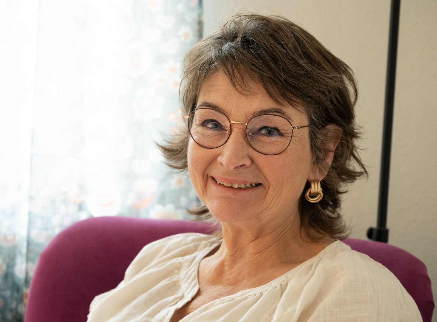 En leende kvinna med kort brunt hår och glasögon sitter i en stol med lila tyg. Hon bär en vit blus och gyllene örhängen. Bakgrunden visar ett ljust fönster med blommigt mönster på gardinen.