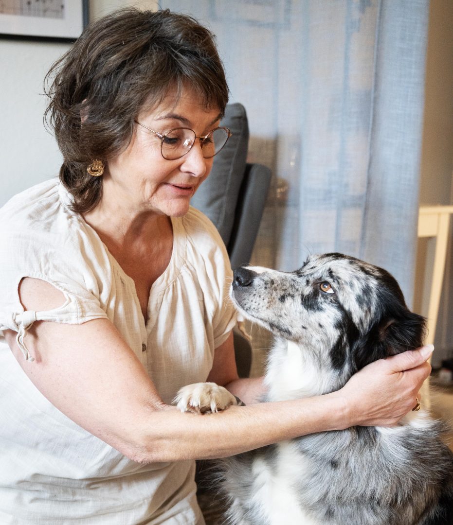 En kvinna med kort brunt hår och glasögon sitter inomhus och håller om en medelstor, långhårig hund med grå, vit och svart päls. Kvinnan ser på hunden med ett varmt uttryck medan hunden tittar tillbaka på henne.