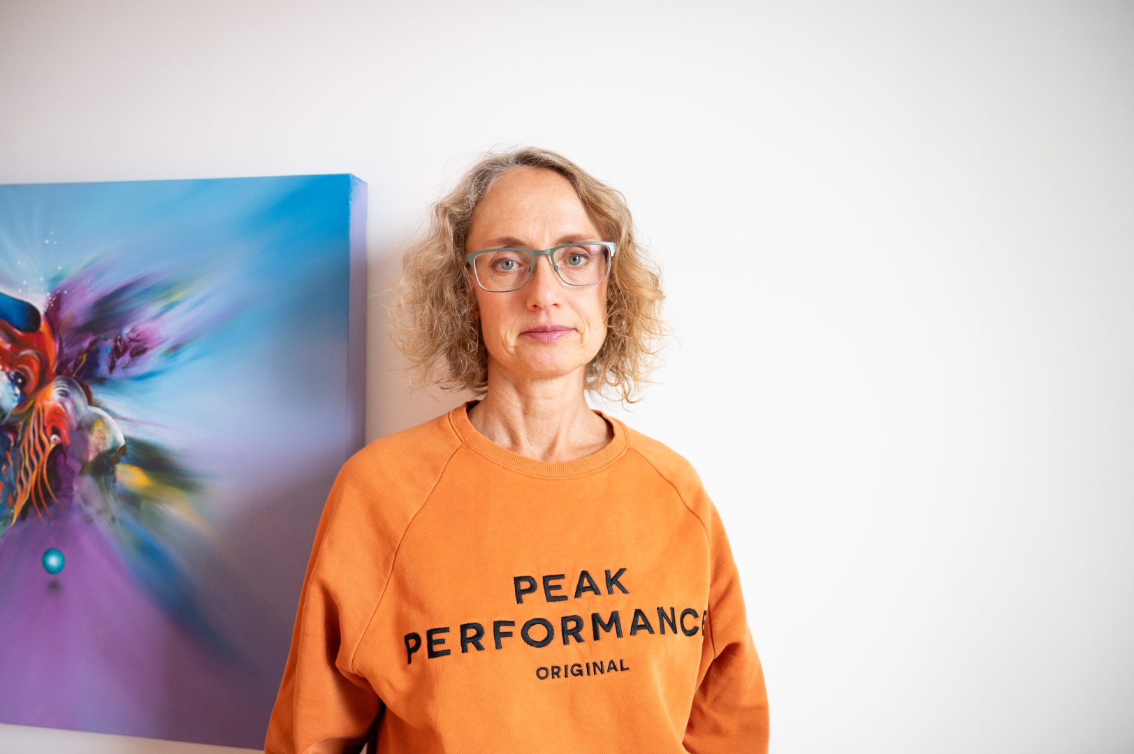 En kvinna med lockigt blont hår och glasögon står mot en vit vägg. Hon bär en orange tröja med texten “Peak Performance Original”. I bakgrunden syns en färgstark tavla i blått och lila.