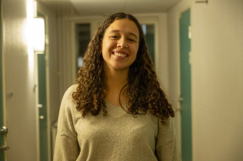 En ung kvinna med lockigt brunt hår står i en upplyst studentkorridor och ler mot kameran. Hon bär en ljusgrå tröja, och dörrar i turkos färg syns längs korridorens väggar i bakgrunden.