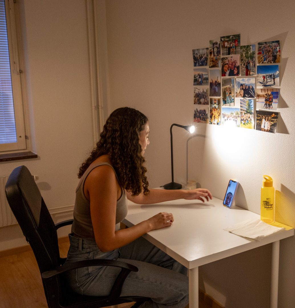 En ung kvinna med lockigt hår sitter vid ett skrivbord i ett studentrum. På väggen framför henne sitter flera färgglada fotografier uppsatta. Hon tittar mot sin mobiltelefon, där ett videosamtal är igång. En skrivbordslampa lyser upp rummet, och bredvid telefonen står en gul vattenflaska.