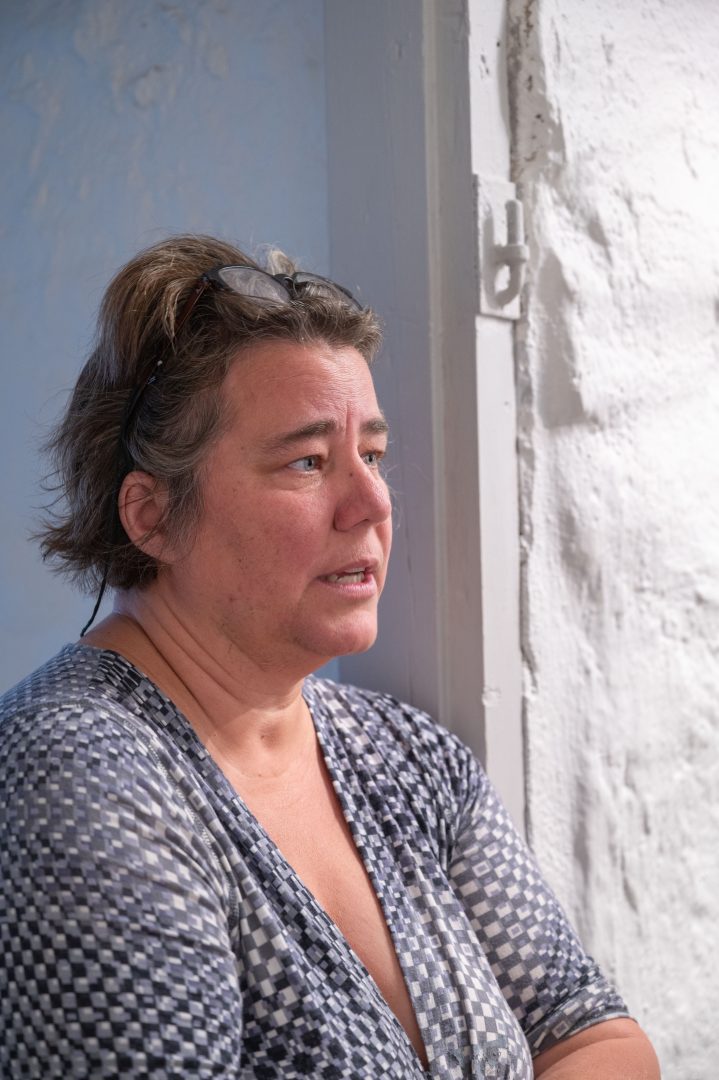 På bilden syns en kvinna som lutar sig mot en vit dörrkarm. Hon har kortklippt hår och glasögon på huvudet. Hon tittar bort från kameran och pratar. Hon har armarna i kors.