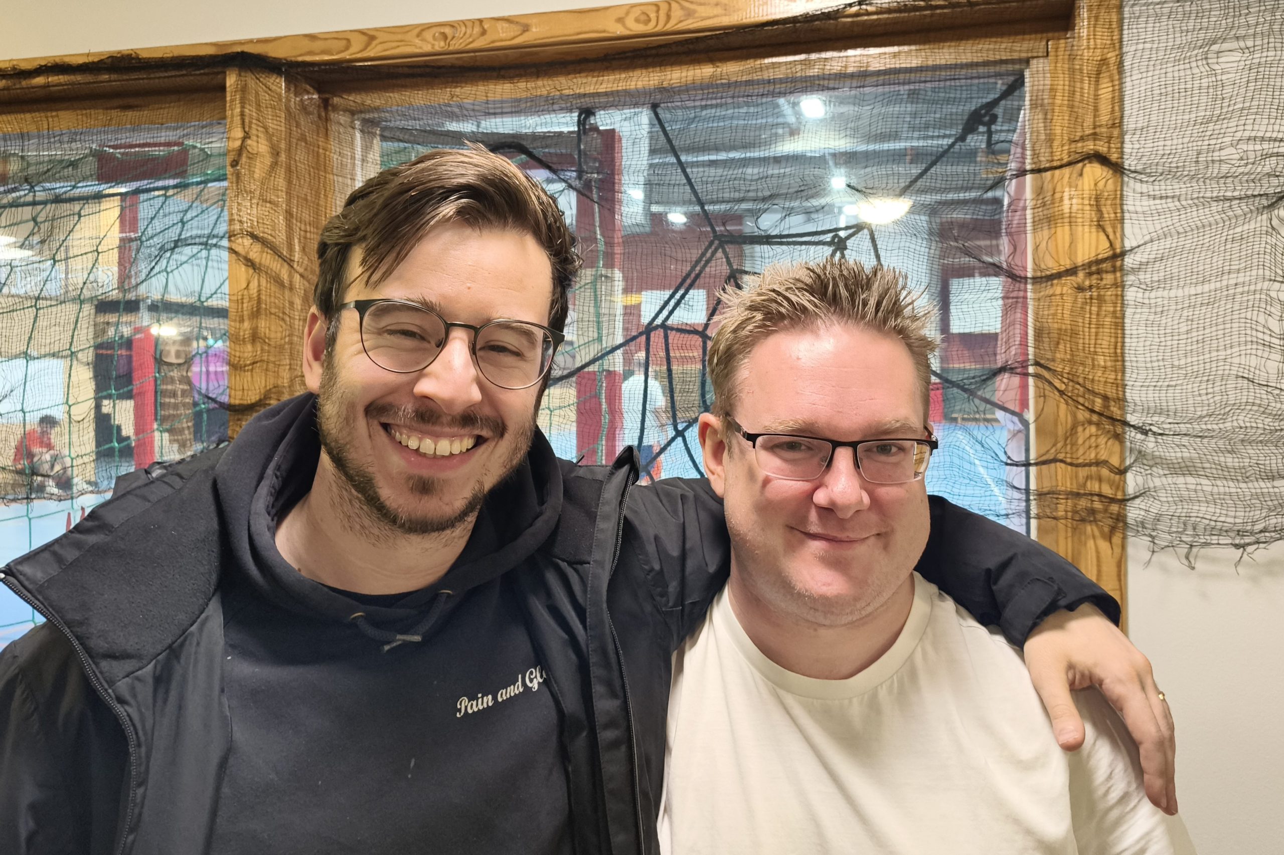 Två män står bredvid varandra inomhus och ler mot kameran. Den ena har mörkt hår och glasögon, den andra har ljust hår och glasögon, och de har en vänskaplig arm om varandras axlar.