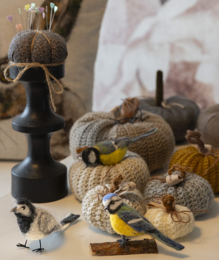 Hantverk står uppradat på ett bord. Nåltovade fåglar, stickade pumpor och en nåldyna av en ljusstake.