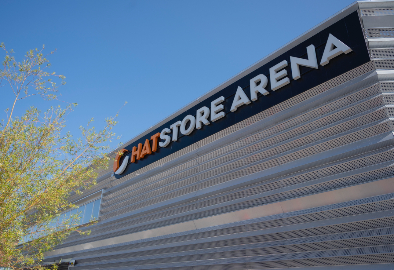 Fasaden på en modern byggnad med metallpaneler och skylten 'HATSTORE ARENA' i stora bokstäver. En grön buske syns till vänster och himlen är klarblå.