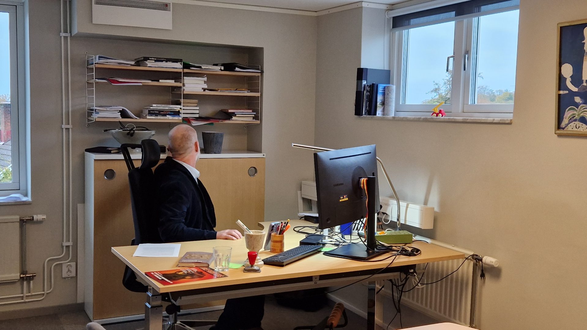 Mannen sitter med ryggen mot kameran vid ett skrivbord inomhus och plockar ut något ur en hylla. Framför honom står en datorskärm och hyllor med böcker syns på väggen.