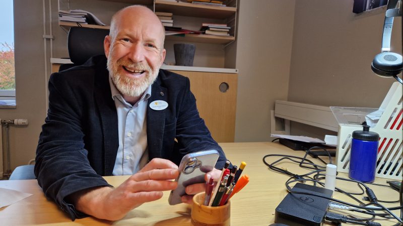 En man sitter vid ett skrivbord inomhus och ler mot kameran medan han håller en mobiltelefon i händerna. På bordet finns pennor, papper och andra kontorsmaterial.