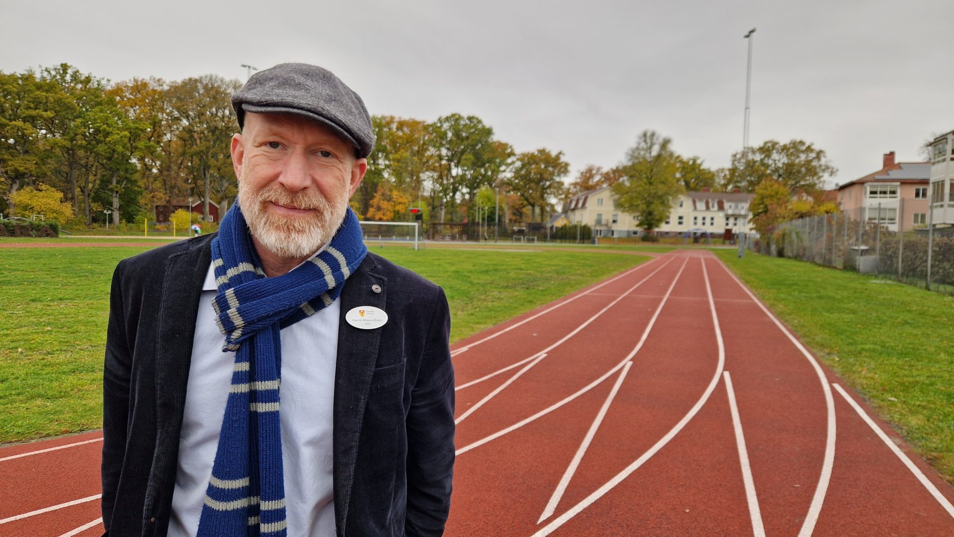 Mannen står på en löparbana utomhus. Han har en grå keps och en blå halsduk på sig. I bakgrunden syns en gräsplan, träd med höstfärger och några byggnader.