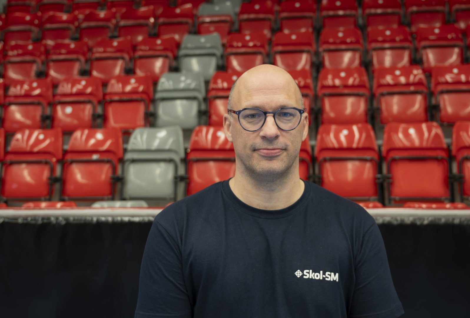 En man i 40 års åldern med svarta glasögon ler framför en läktare av röda stolar