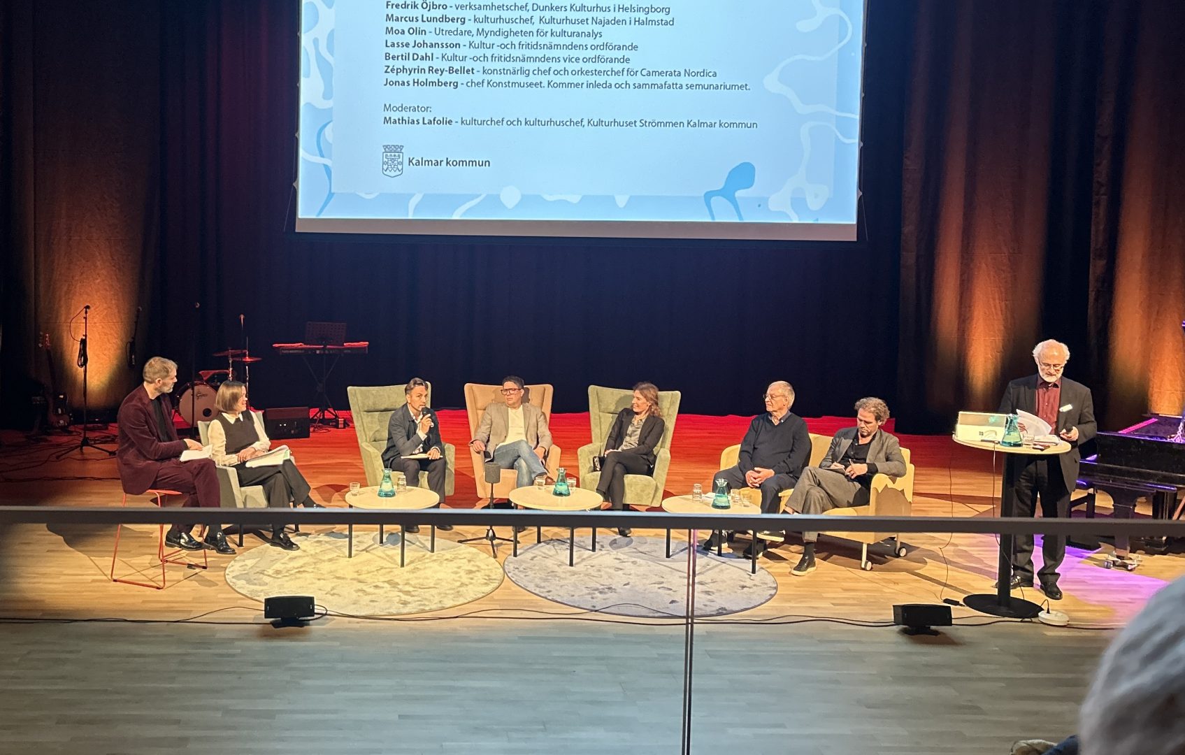 Bilden visar en paneldiskussion eller intervju på en scen. Det finns sju personer, fem sittande i fåtöljer och två står eller sitter vid små bord eller podier. Bakom dem syns ett stort skärmar med text och logga, samt några musikinstrument. Scenen är upplyst med varma färger och har ett mörkt bakgrundstält.