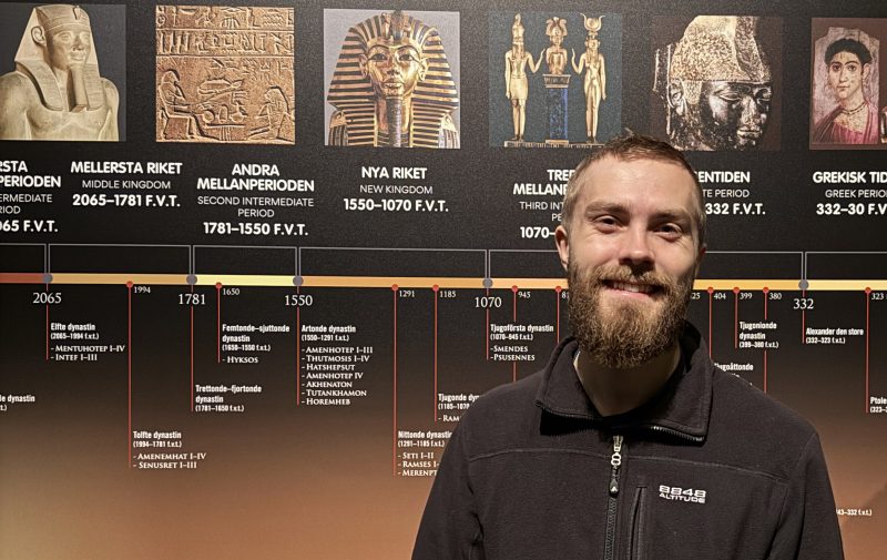 En person står framför en väggutställning som visar en tidslinje över forntida Egyptens olika historiska perioder. Tidslinjen är illustrerad med bilder på faraoner, hieroglyfer, statyer och målningar från de olika epokerna.