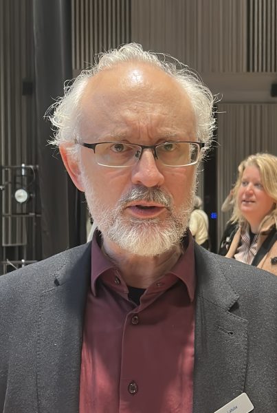 Bilden visar en man med grått hår och skägg som bär glasögon. Han har på sig en mörkröd skjorta och en mörk kavaj. Bakom honom syns en kvinna och en inomhusmiljö med lodräta väggpaneler och dämpad belysning.