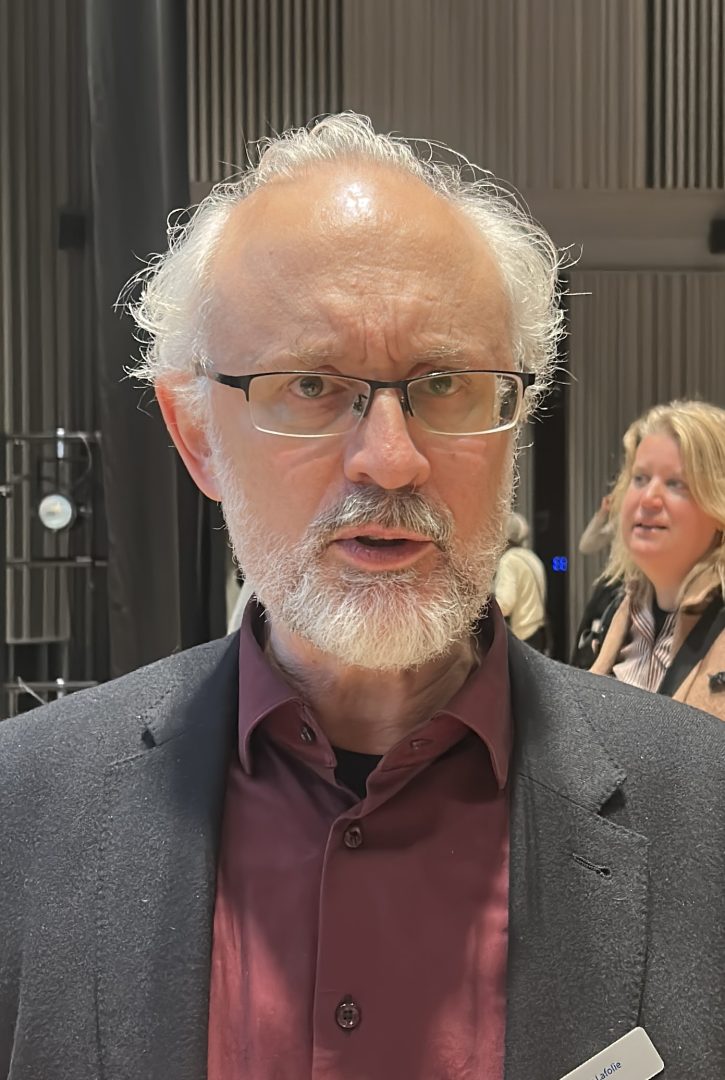 Bilden visar en man med grått hår och skägg som bär glasögon. Han har på sig en mörkröd skjorta och en mörk kavaj. Bakom honom syns en kvinna och en inomhusmiljö med lodräta väggpaneler och dämpad belysning.