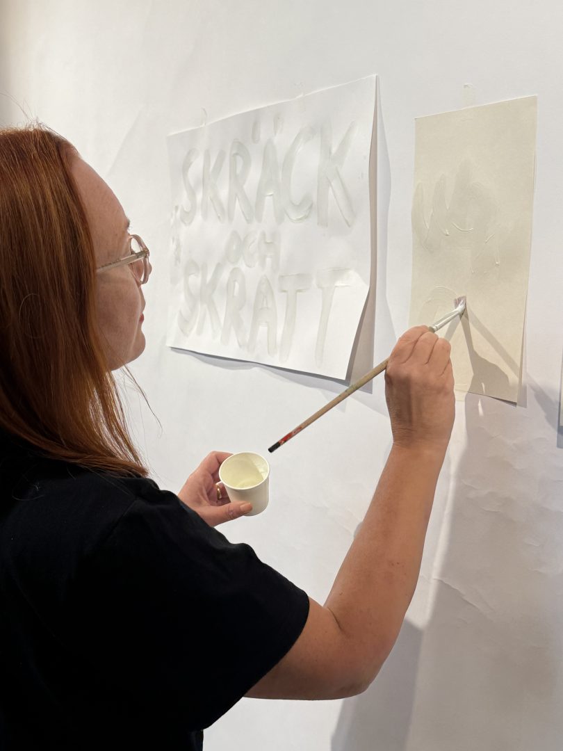 En person med rött hår och glasögon målar med pensel på ett vitt papper som sitter upptejpat på väggen. På pappret står texten ”Skräck och skratt” målad i vit färg. Personen håller en liten kopp med färg i den andra handen.