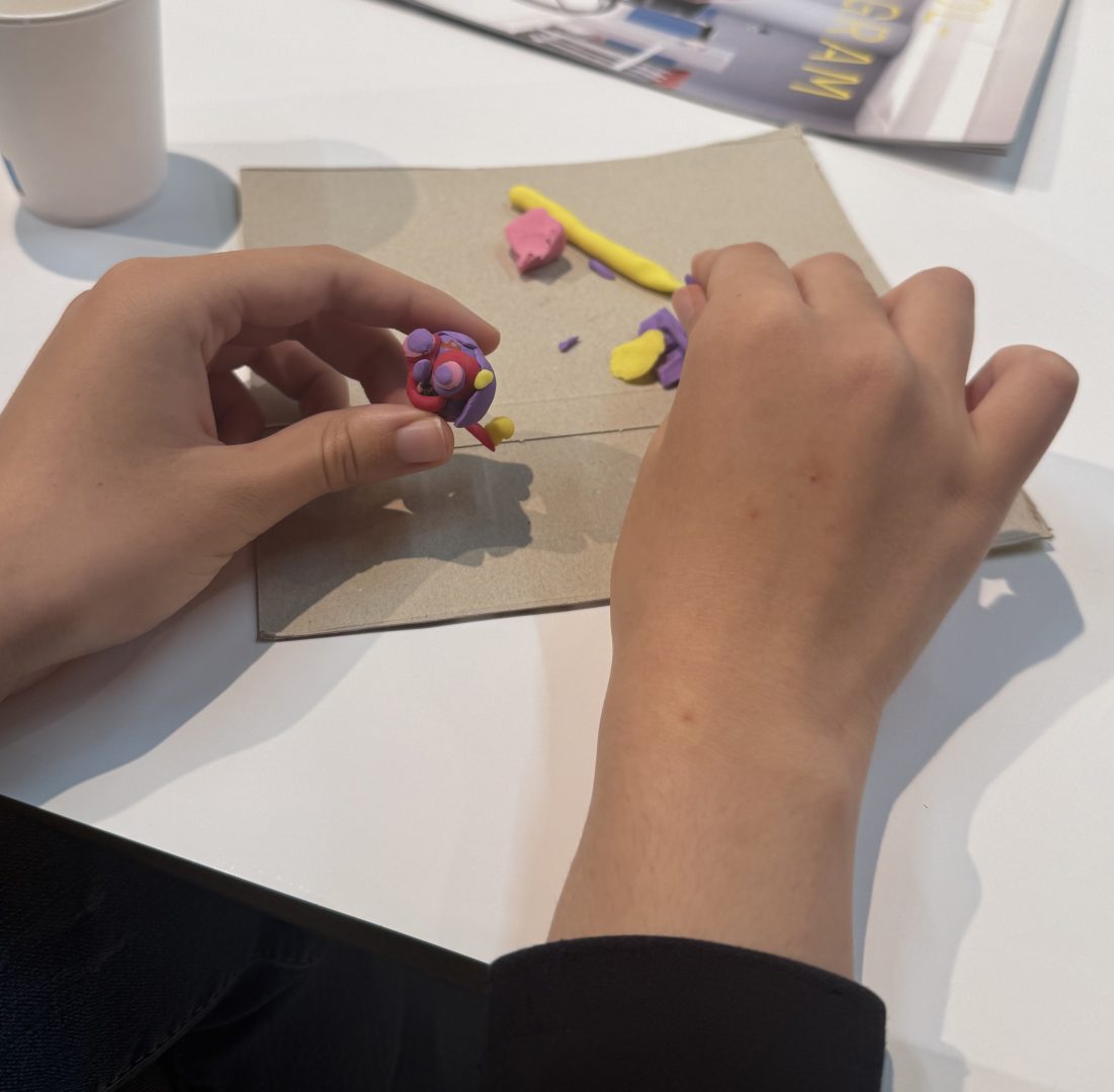 Ett par händer formar en liten figur av färgad lera i rosa, lila och gult. Figuren hålls över en bit kartong på ett bord där fler bitar av lera ligger utspridda. I bakgrunden syns en pappersmugg och en tidskrift.