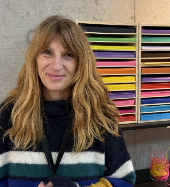 En kvinna med långt, ljusbrunt hår står framför en hylla fylld med färgglada pappersark i olika nyanser. Hon bär en stickad tröja med breda ränder i blått, vitt, grönt och gult, och ler lätt mot kameran. Bakgrunden består av en grå vägg och en bänk med en burk fylld med färgglada snören.