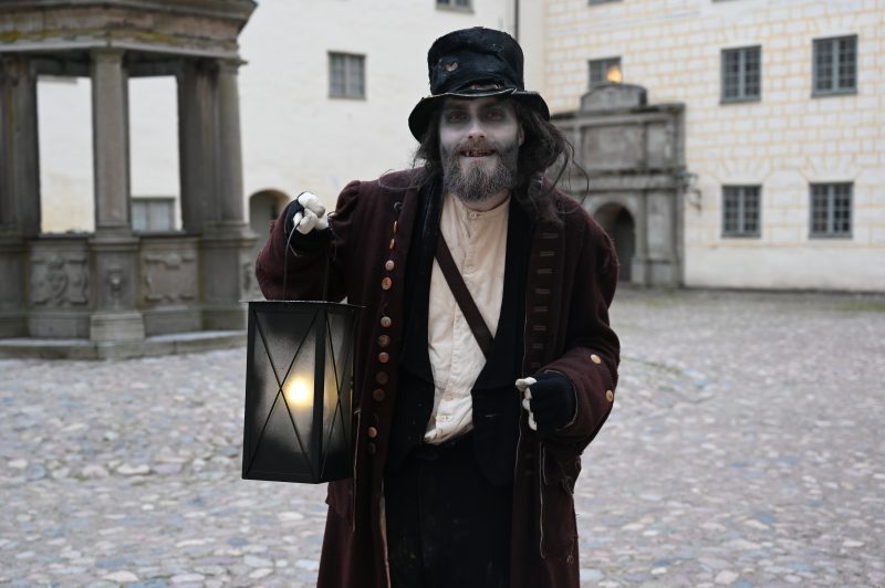 En person utklädd till en spöklik figur med vitmålat ansikte, rufsigt hår och skägg, klädd i mörk, gammaldags rock och hatt, håller en tänd lykta. Bakgrunden visar en borggård med stenbeläggning och gamla byggnader.