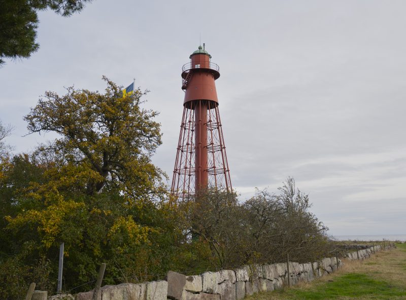 En hög röd metallfyr står på en gräsbevuxen plats vid kusten. Framför fyren syns träd med gula höstlöv, en stenmur och en svensk flagga som sticker upp bakom träden. Himlen är grå och havet skymtar i bakgrunden.