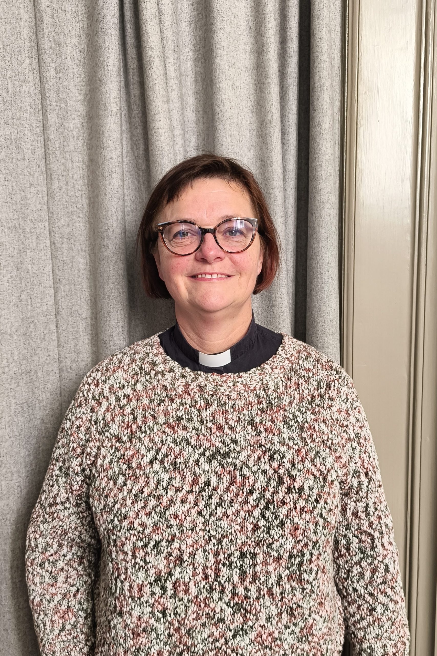 En person står framför ett grått draperi och en ljus vägg. Hen bär glasögon, en mönstrad stickad tröja och en prästskjorta med vit krage, och ler mot kameran.