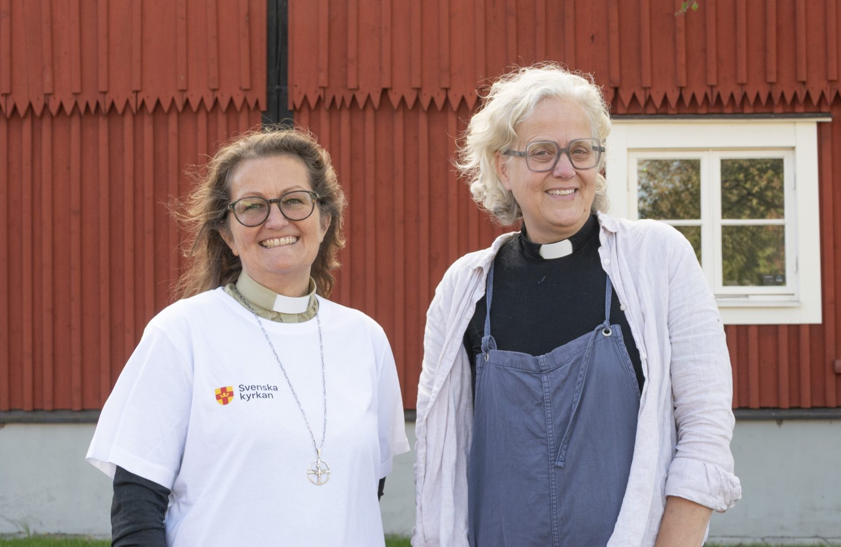Linda Ursjö, diakon och Sofia Ekelund Fogelström, präst i Förlösa Kläckeberga församling ler framför den röda träbyggnaden Garvaregården i Lindsdal.