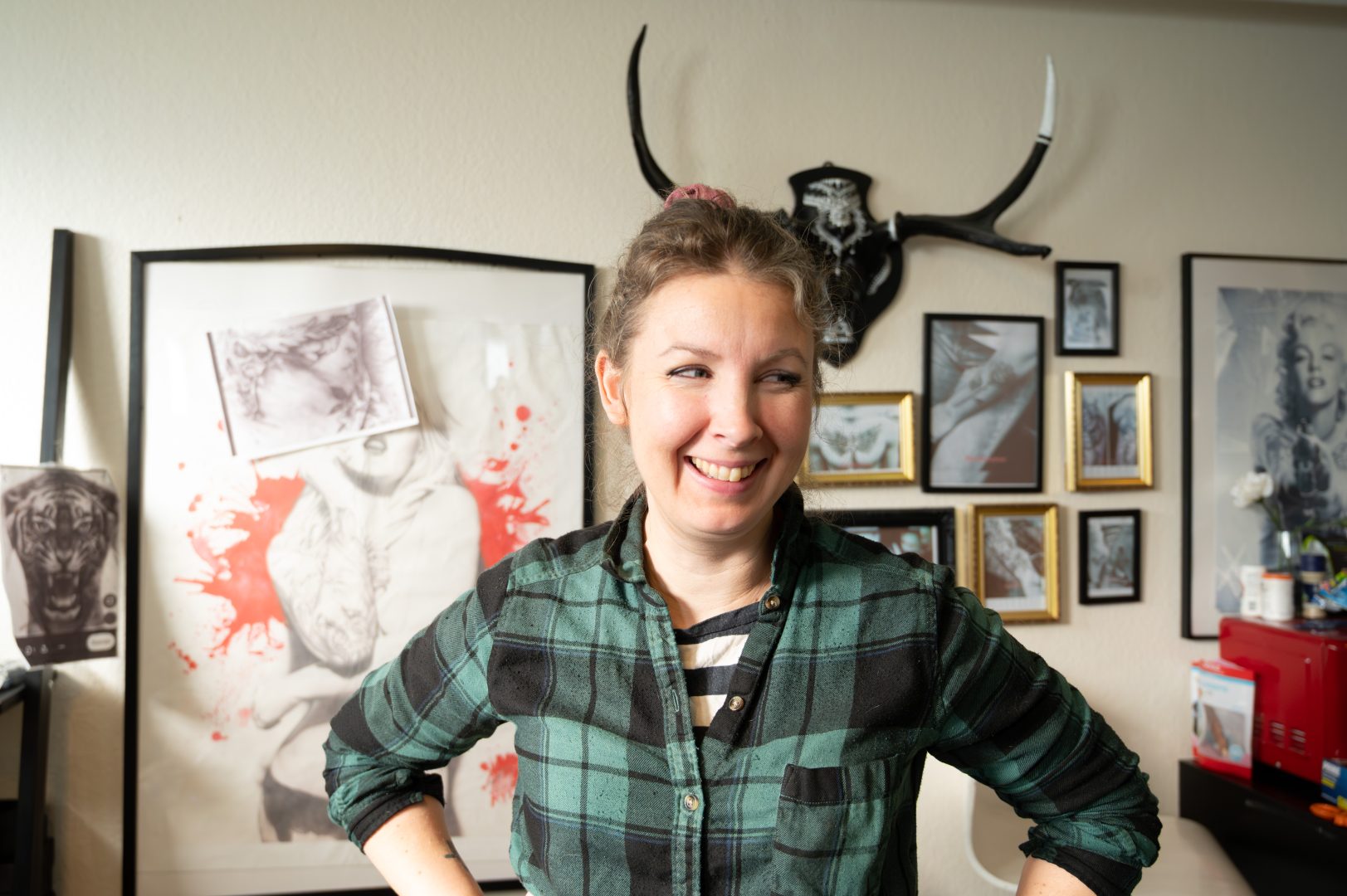 En person står inomhus och ler. Hen har uppsatt hår och bär en grön- och svartmönstrad flanellskjorta. I bakgrunden syns flera konstverk på väggen, bland annat teckningar och målningar i svart, vitt och rött. Där finns också en väggprydnad med horn samt några inramade bilder i olika storlekar. Ljuset i rummet är varmt och naturligt.